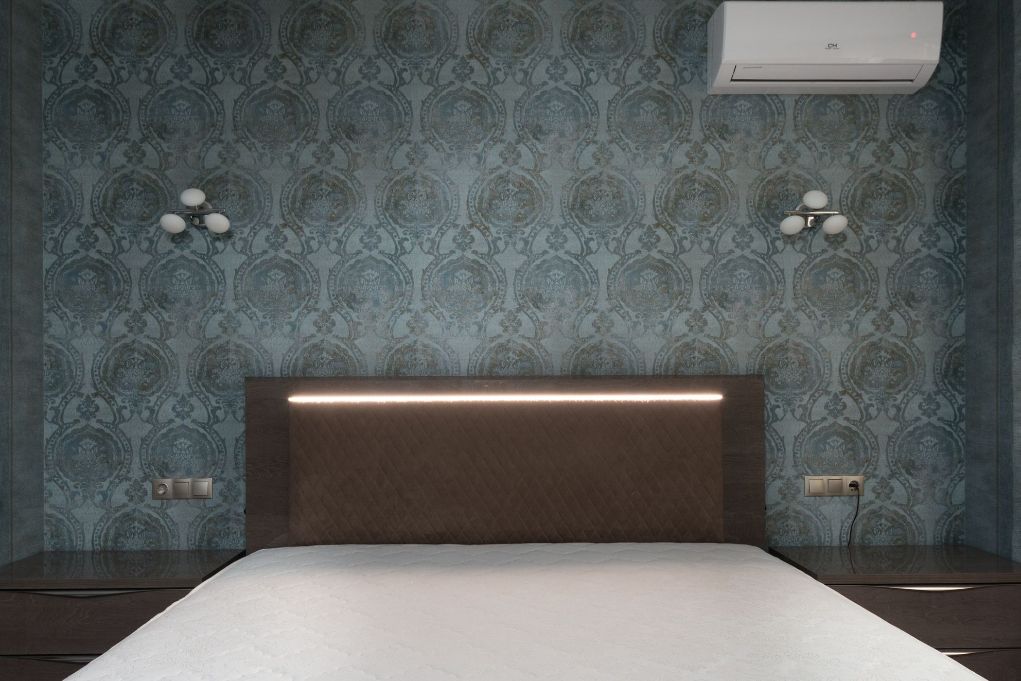 Ornamental textured wallpaper behind a wooden headboard and white wall lamps
